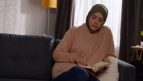 Mujer-Musulmana-Con-Hijab-Sentada-En-Un-Sofá-En-Casa-Leyendo-O-Estudiando-El-Corán-2
